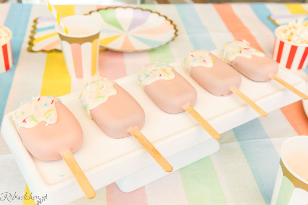 Cakesicles liegen auf einer Platte auf dem Tisch und sind mit Glasur und Streuseln verziert